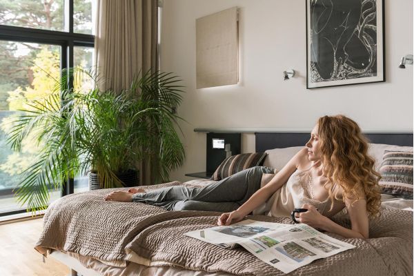 Crestron control panel in a bedroom and a woman reading a newspaper and drinking coffee layed in bed