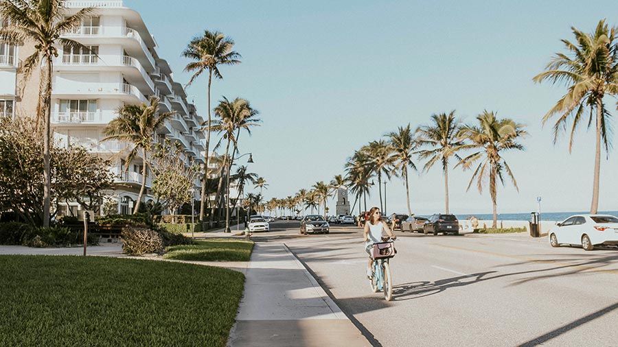 Woman biking along a coastal road with palm trees and condos